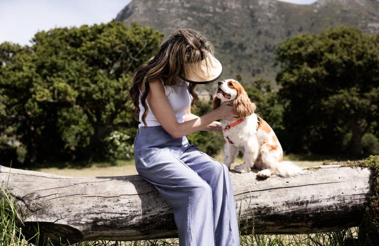 Brown haired women with a visor and brown and white cavalier sitting on a log in the sunshine