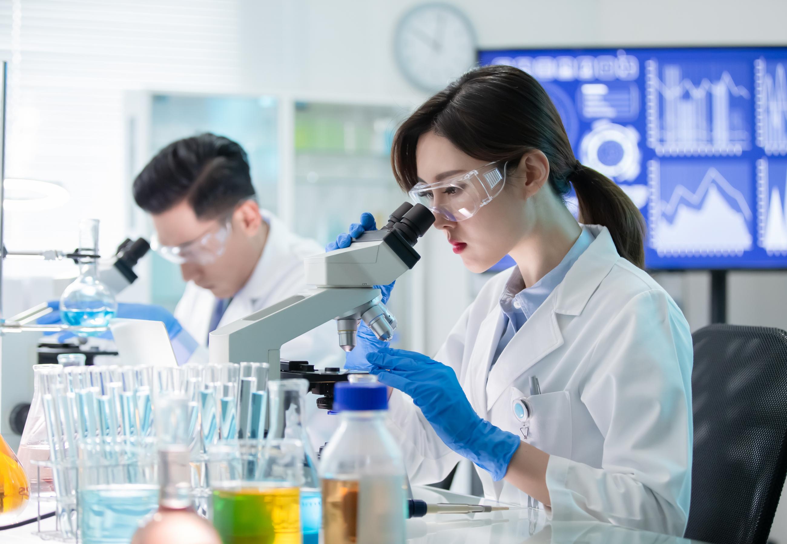 Two scientists wearing goggles sitting at microscopes in a laboratory